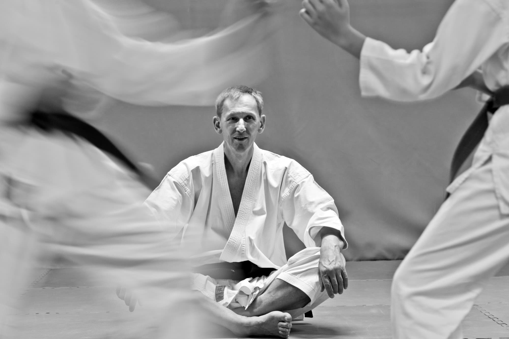 Czarno-białe zdjęcie: sensei Mirosław Ellwart, uśmiechnięty, w karategi z czarnym pasem, siedzi po turecku na macie, a przed nim, rozmyte w ruchu, przebiegają dwie osoby w karategi