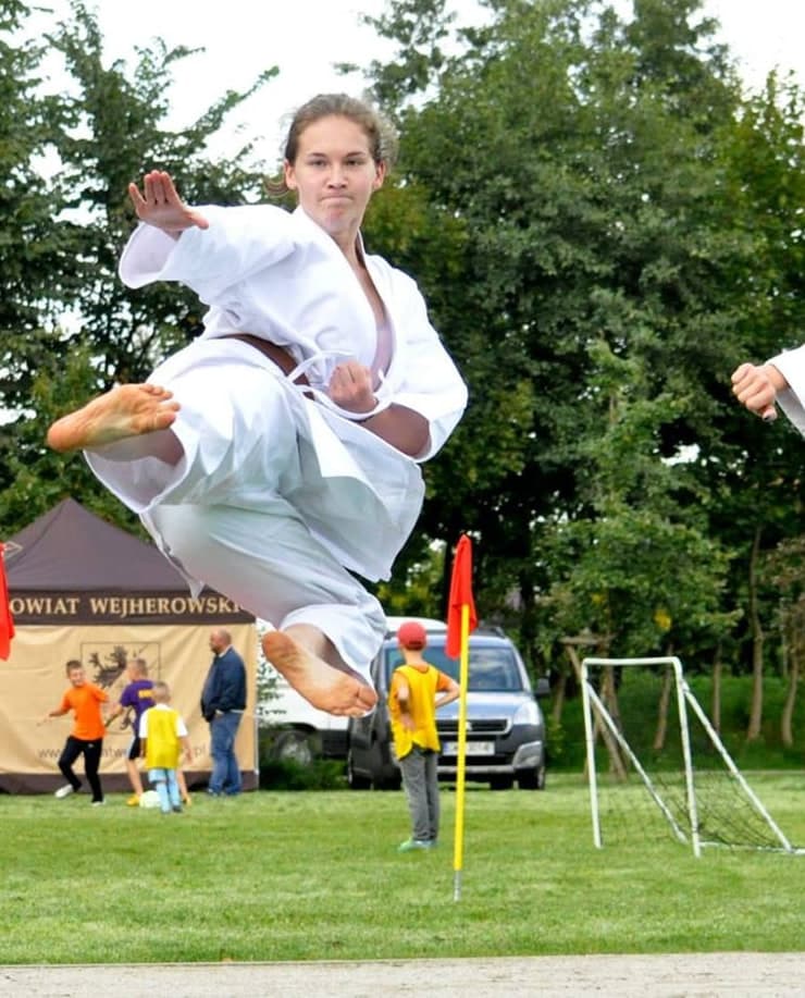Zawodniczka w białym karategi z brązowym pasem w locie, w trakcie kopnięcia w wyskoku podczas plenerowego pokazu karate na trawiastym boisku; w tle drzewa, baner Powiatu Wejherowskiego i publiczność