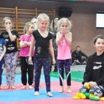 Grupa przedszkolaków w kolorowych strojach sportowych stoi w rzędzie na macie podczas konkurencji, obok instruktorka w czarnym stroju