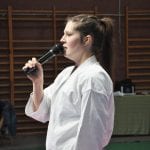 Instruktorka w karategi mówi do mikrofonu podczas zawodów, w tle drabinki hali