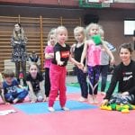 Dzieci w strojach sportowych czekają na swoją kolej na czerwono-niebieskiej macie, z prawej instruktorka pilnująca przebiegu konkurencji