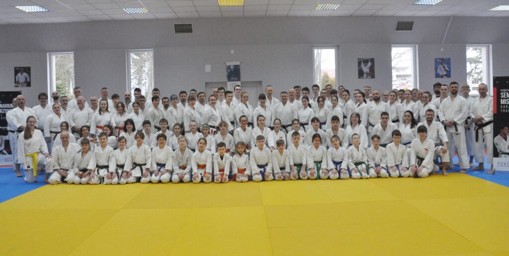 Ponad stuosobowa grupa uczestników seminarium w karategi pozuje w kilku rzędach w jasnej sali, przed nimi żółta mata, na ścianach portrety mistrzów karate