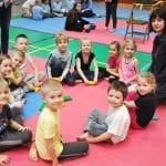 Grupa przedszkolaków siedzi na macie w kilku rzędach i pozuje do zdjęcia, obok opiekunki