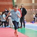 Chłopiec w niebieskiej koszulce biegnie po torze przeszkód na hali, w tle sędzia i publiczność
