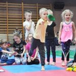 Dzieci w strojach sportowych wykonują ćwiczenie z woreczkiem na macie, obok siedzą pozostali uczestnicy
