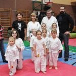 Drużyna przedszkolaków w karategi i strojach sportowych pozuje z instruktorami i opiekunami na macie hali