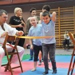Chłopiec przechodzi pod rozciągniętą taśmą w konkurencji sprawnościowej, obok instruktor w karategi i opiekunowie