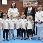 Drużyna przedszkolaków w białych koszulkach pozuje z opiekunkami i instruktorką w karategi na macie hali