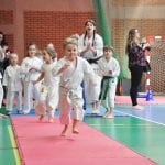 Dziewczynka w karategi biegnie po czerwonej macie, za nią kolejni zawodnicy i instruktorzy