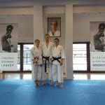 Troje karateków z czarnymi pasami stoi obok siebie na macie w dojo, pod portretami mistrzów karate tradycyjnego