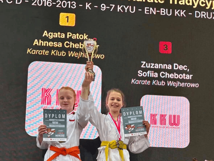 Dwie dziewczynki w karategi z pomarańczowym i żółtym pasem na podium: jedna trzyma uniesiony puchar, obie pokazują dyplomy, za nimi ekran z wynikiem drużynowym Karate Klubu Wejherowo