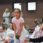 Dziewczynka w różowej koszulce wykonuje ćwiczenie na macie, wokół siedzą pozostali uczestnicy, w tle opiekunka