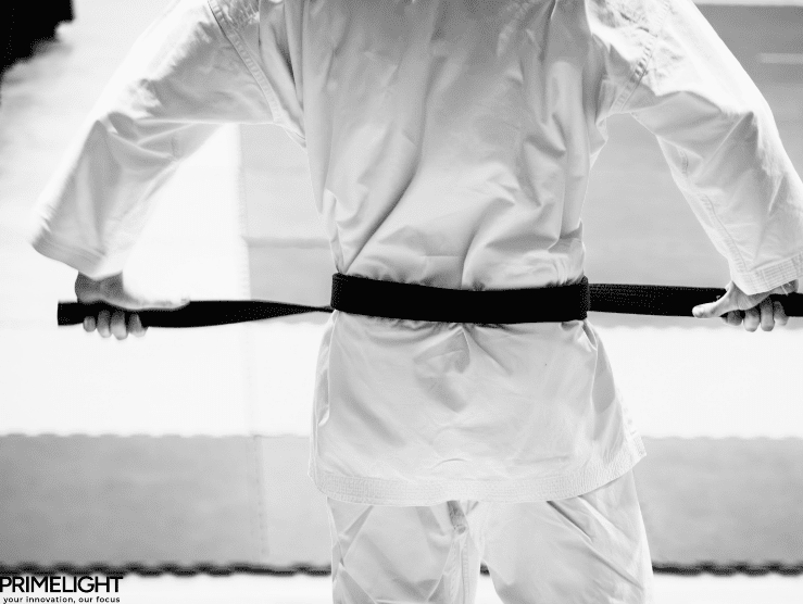Czarno-białe zdjęcie: karateka widziany od tyłu trzyma rozciągnięty w rękach czarny pas na wysokości bioder, stojąc na macie