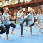 Grupa dzieci w błękitnych koszulkach ćwiczy uderzenie w wykroku na niebieskiej macie hali