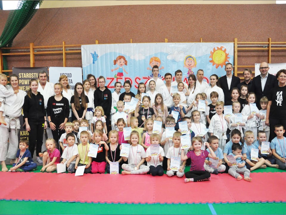 Pamiątkowe zdjęcie wszystkich uczestników Przedszkolandii: kilkadziesiąt dzieci z dyplomami i medalami siedzi i stoi przed banerem imprezy, za nimi opiekunowie, instruktorzy w karategi i goście, po bokach banery Starostwa Powiatowego i Urzędu Miasta