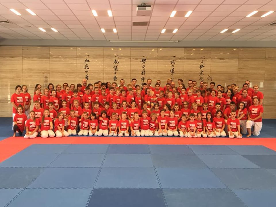 Blisko stu uczestników obozu w czerwonych koszulkach klubowych i białych spodniach karategi pozuje w kilku rzędach na niebiesko-czerwonej macie dojo, na ścianie japońska kaligrafia zasad dojo kun