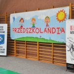 Baner Przedszkolandii z rysunkami dzieci i słońcem rozwieszony na drabinkach hali sportowej