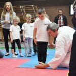 Instruktor kuca przy macie i ustawia przeszkodę, obok czekają dzieci w białych koszulkach