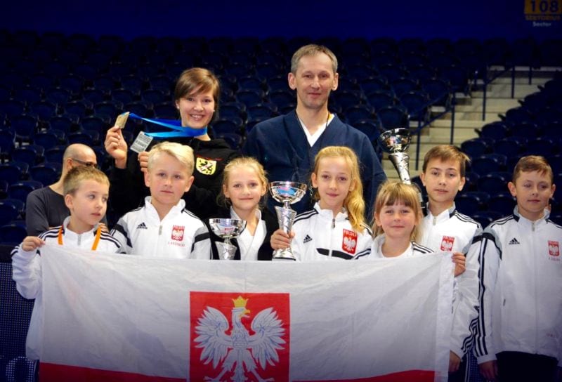 Dzieci w białych dresach reprezentacji Polski trzymają biało-czerwoną flagę i puchary, za nimi trenerka z medalem i trener w granatowym hakama; w tle puste trybuny hali w Wilnie