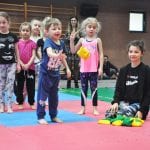 Dzieci w strojach sportowych stoją na macie przed konkurencją z kolorowymi przyborami rozłożonymi obok