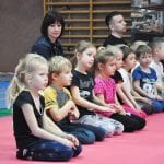 Dzieci siedzą po turecku na czerwonej macie, za nimi opiekunowie obserwujący ceremonię