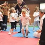Mała dziewczynka w różowej koszulce idzie po macie w konkurencji, obok opiekunowie i pozostałe dzieci