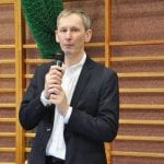 Mężczyzna w ciemnej marynarce przemawia do mikrofonu w hali, w tle drabinki i zielona siatka