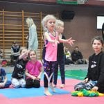 Dziewczynka w różowej koszulce stoi na macie w trakcie konkurencji, wokół siedzą uczestnicy i opiekunka