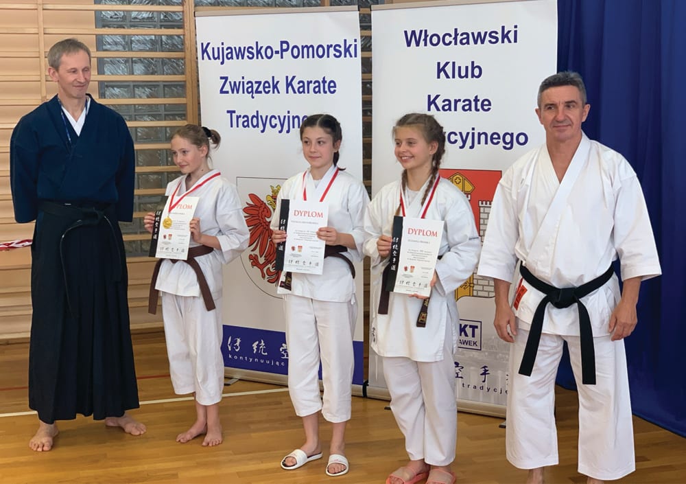 Trzy zawodniczki z brązowymi pasami trzymają dyplomy i medale, po bokach dwaj trenerzy - jeden w granatowym hakama, drugi w karategi z czarnym pasem; w tle banery Kujawsko-Pomorskiego Związku Karate Tradycyjnego i Włocławskiego Klubu Karate Tradycyjnego