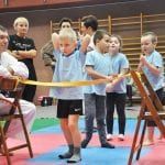 Dzieci czekają w kolejce przed przeszkodą z rozciągniętą taśmą, obok instruktor i opiekunowie