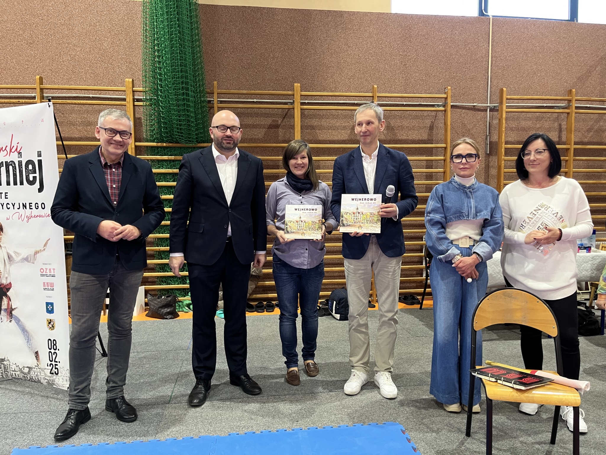 Sześć osób pozuje w hali sportowej przed banerem Pomorskiego Turnieju Karate Tradycyjnego z datą 08.02.'25; dwoje z nich trzyma albumy o Wejherowie, w tle drabinki gimnastyczne